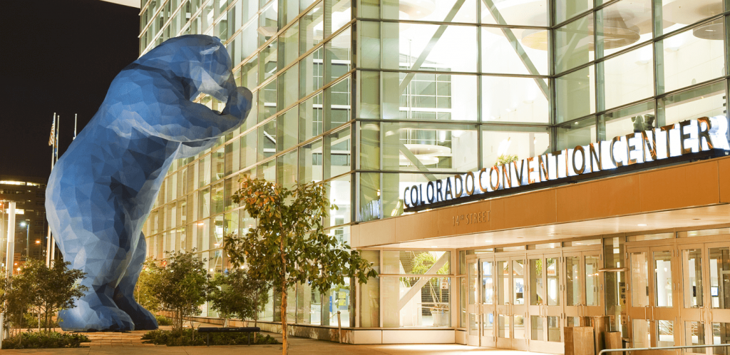 Denver's iconic blue bear sculpture peering into the windows of the Colorado Convention Center at night, with the building's glass facade illuminated behind it.
