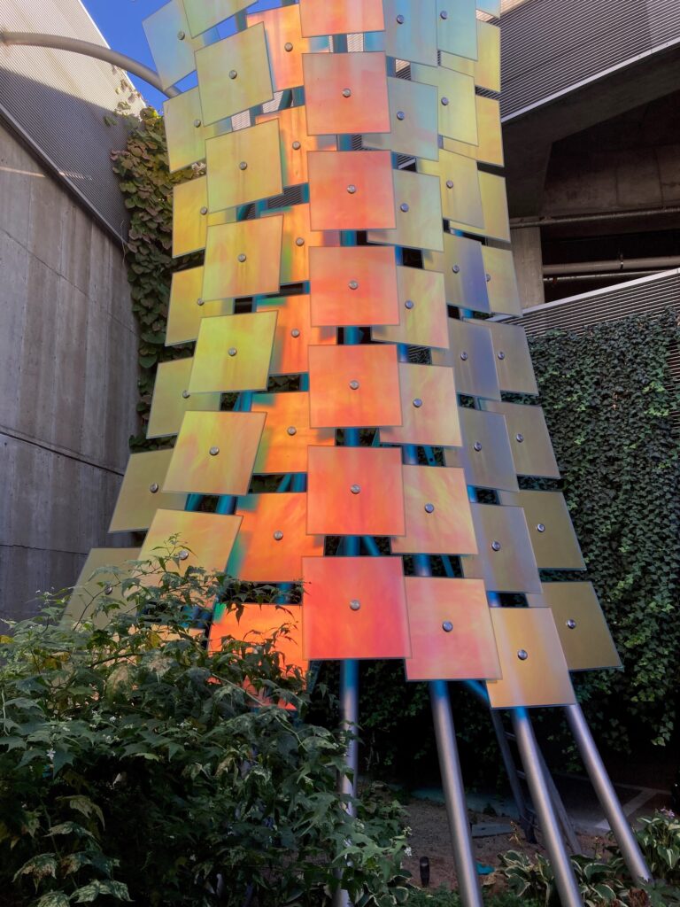 A tall tower sculpture covered in overlapping iridescent square glass panels that shift between orange, yellow, pink, and blue depending on the light. The piece is mounted on steel poles in an outdoor courtyard surrounded by ivy-covered walls and green shrubs.
