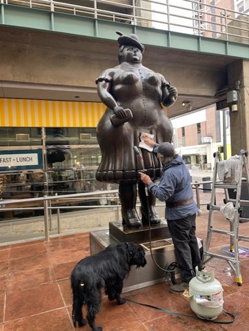 A conservator works on one of the large bronze Botero sculptures at the Denver Performing Arts Complex, cleaning the surface with a cloth. A black dog stands nearby. A propane tank and ladder are visible beside the sculpture.