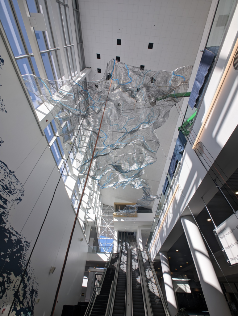 An interior view looking up at the large suspended sculpture at the Colorado Convention Center, photographed during installation. The billowing metallic mesh is printed with black topographic contour lines and blue threads suggesting rivers, and hangs from the high ceiling above a pair of escalators. Installation rigging and ropes are still visible.