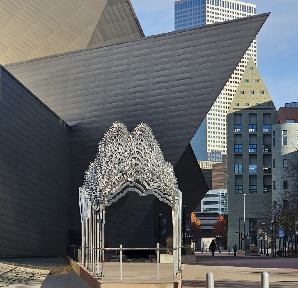 A large walk-through sculpture made of stacked polished steel arches forming a gateway structure, installed outside the Denver Art Museum. The repeating arch forms suggest mountain peaks or cresting waves. The angular black titanium facade of the museum and downtown Denver skyscrapers are visible behind it under a clear blue sky.