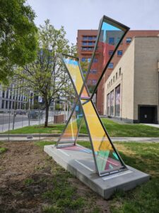 A large X-shaped sculpture made of stainless steel and dichroic glass, installed on a concrete base outside the Denver Central Library near West 14th Ave. and Broadway. The two crossing panels of glass shift between yellow, pink, blue, and teal depending on the light and viewing angle. The sculpture faces the State Capitol building. Spring trees, a red brick building, and the library's stone facade are visible in the background.