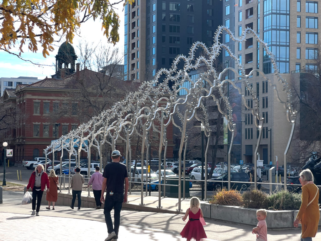 A street-level view of the 'Trestle' sculpture with pedestrians and children passing by on a sunny autumn day. The polished steel arched structure stretches along the plaza, with a historic red brick building, bare trees, and downtown high-rises visible in the background.