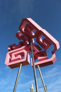 A close-up view looking up at a large pink and red geometric sculpture mounted on steel poles against a deep blue sky. The piece features interlocking rectangular forms with spiral and maze-like cutouts, creating a layered, three-dimensional composition. The Denver City and County Building clock tower is partially visible at the base of the poles.