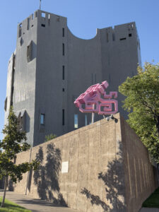 A wide shot of the pink geometric 'Glyph' sculpture mounted on poles in front of the Denver Public Library's brutalist gray stone tower. The bold, maze-like form contrasts with the angular architecture behind it. Trees and sidewalk are visible at the base.