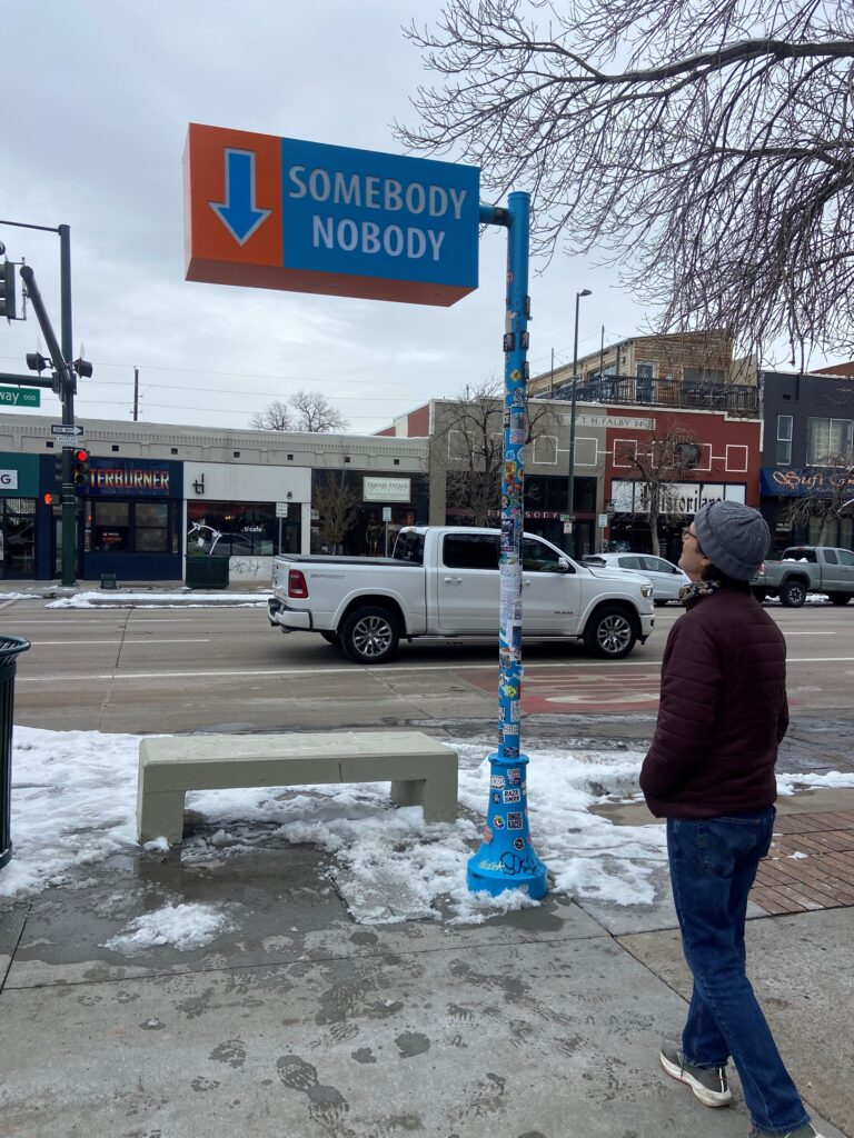 A person stands on a snowy sidewalk looking up at a street-style sign mounted on a bright blue pole covered in stickers. The sign features a blue downward-pointing arrow on an orange background alongside a blue panel reading 'Somebody Nobody' in white text. A concrete bench sits nearby. The surrounding South Broadway streetscape includes storefronts, bare winter trees, and passing traffic under an overcast sky.