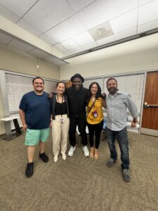 Five people stand together smiling in an office meeting room, posing for a group photo.