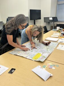 Two people lean over a large printed map spread across a conference table, with one pointing to a location. Colored dot stickers and papers are scattered on the table nearby.
