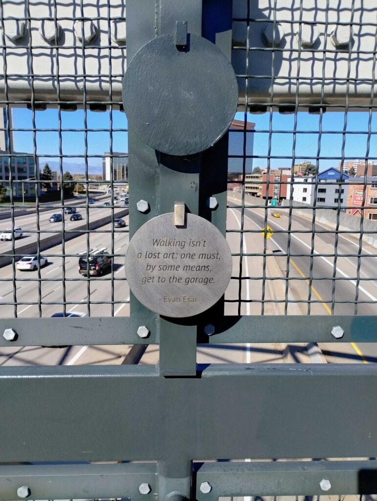 A close-up of a circular metal plaque mounted on the steel mesh railing of a pedestrian bridge. The plaque reads: 'Walking isn't a lost art: one must, by some means, get to the garage. – Evan Esar.' A busy highway with moving cars is visible through the mesh below, with Denver city buildings and the Rocky Mountains in the background under a clear blue sky.