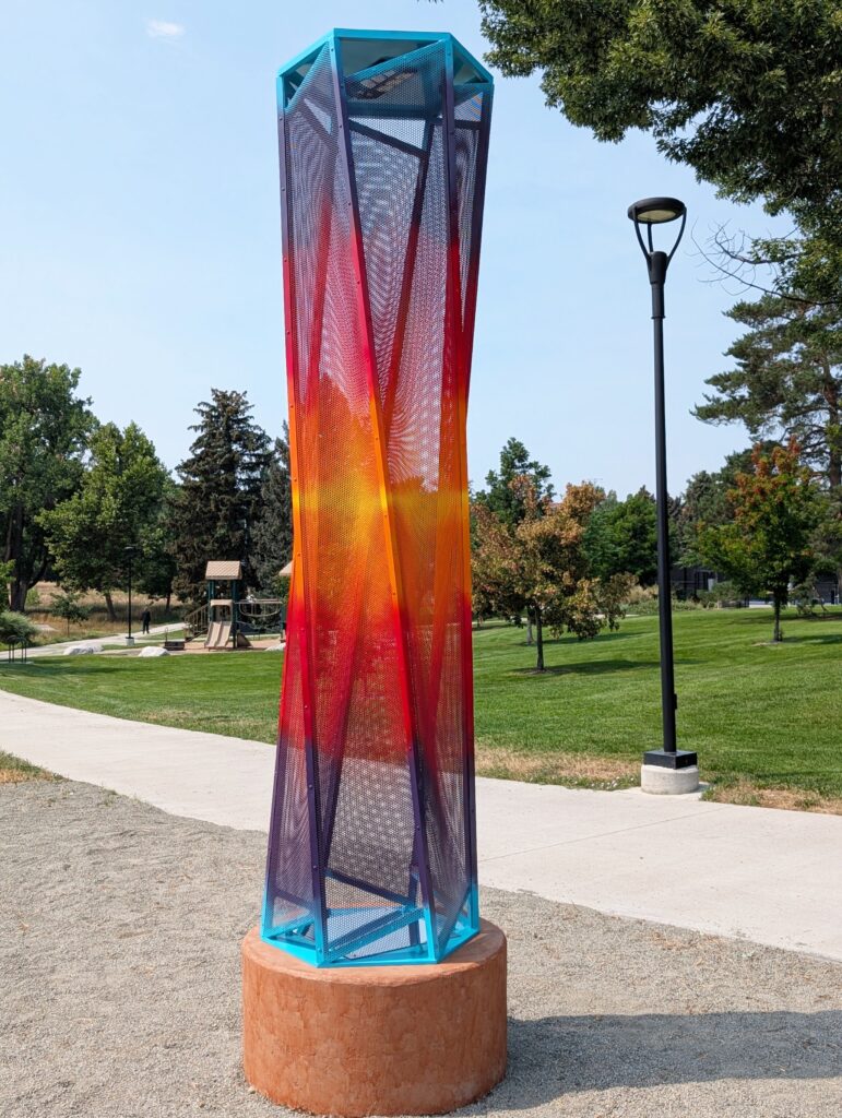 A tall, twisting hexagonal sculpture on a terracotta-colored concrete base in Bear Valley Park. The structure is made of perforated mesh panels that spiral gently from base to top, with colors gradating from deep purple at the bottom through red and orange to bright cyan at the top edges and cap. Trees, a playground, and a park path are visible in the background on a sunny day.