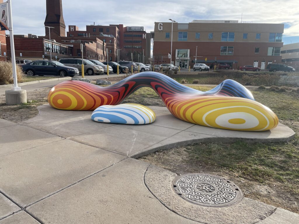A large, biomorphic sculpture on a concrete pad outside the Denver Health campus. The piece consists of two rounded, lobed forms connected by a curving bridge, covered in bold concentric stripe patterns. The left form graduates from orange and red to dark tones at the arch; the right form features yellow and white concentric circles. A smaller, separate rounded form in light blue and white stripes sits in front. Hospital buildings and a parking lot are visible in the background.