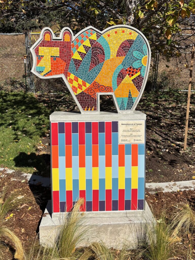 A mosaic sculpture of a stylized bear on a colorful tiled pedestal at Joe Martinez Park. The bear form faces left and is filled with bold geometric mosaic patterns in teal, red, orange, yellow, and maroon, with a smaller figure nestled against its back. The matching tiled pedestal features vertical stripes of red, orange, yellow, blue-gray, and purple. A bilingual artwork plaque is mounted on the front of the pedestal.