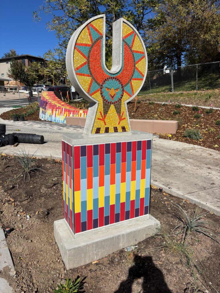 A mosaic sculpture of a stylized eagle on a colorful tiled pedestal at Joe Martinez Park. The eagle is depicted in a circular sunburst form, with red, teal, and yellow mosaic tiles forming radiating feathers and a bird face at the center. The pedestal is covered in a grid of vertical rectangular tiles in red, orange, yellow, blue-gray, and purple. A colorful mosaic wall is partially visible in the background.