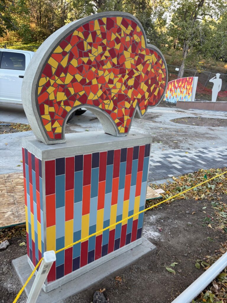 A mosaic sculpture of a stylized bison on a colorful tiled pedestal at Joe Martinez Park, photographed during installation. The bison silhouette is filled with large irregular mosaic fragments in red, orange, and yellow. The pedestal features the series' signature vertical striped tile pattern in warm and cool tones. Yellow caution tape and construction materials are visible in the background, along with another sculpture in the series.