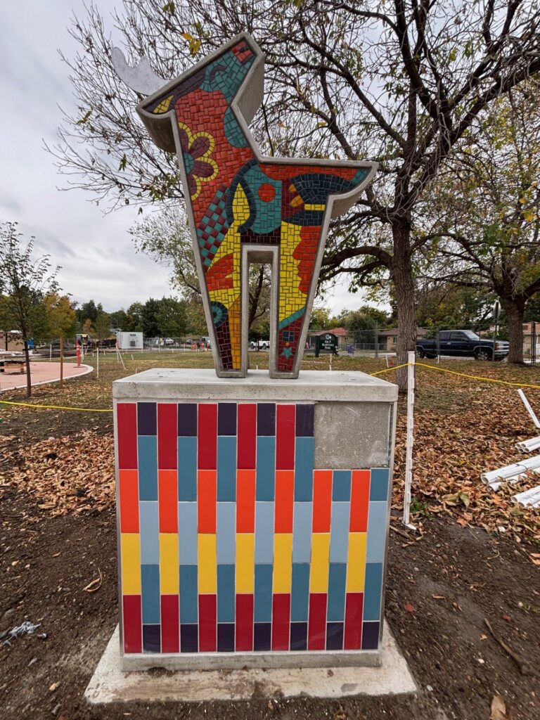 A mosaic sculpture of a stylized deer on a colorful tiled pedestal at Joe Martinez Park, photographed during installation. The deer form faces left with abstract mosaic patterns in red, teal, yellow, and maroon. The pedestal matches the series' striped tile pattern. A park sign and construction materials are visible in the background.