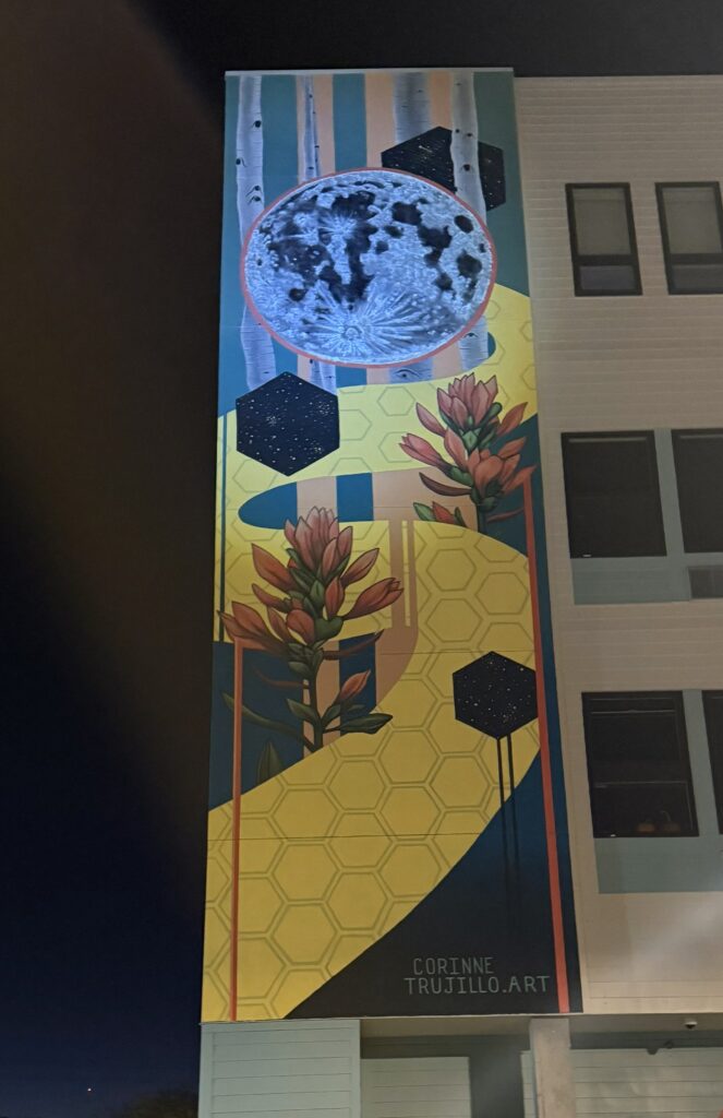 A tall, narrow exterior mural on the side of a multi-story building, photographed at night. The composition features a large detailed moon at the top surrounded by aspen trees, floating dark geometric cube forms dusted with stars, two Indian paintbrush wildflowers, and a sweeping yellow honeycomb-patterned band running diagonally through the center against a teal background. The artist's signature 'Corinne Trujillo.Art' is visible at the bottom.