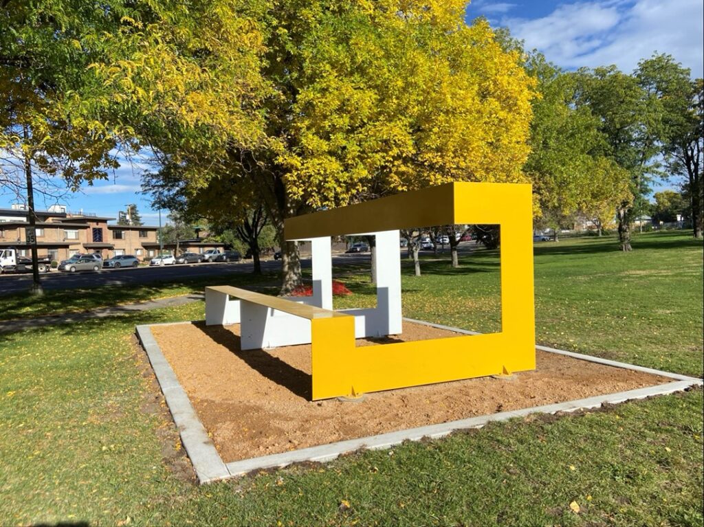 A bright yellow rectangular steel frame sculpture enclosing a white bench, installed on a gravel pad at Burns Park. The open geometric form resembles a large picture frame or portal. Autumn trees in yellow and green foliage frame the background under a partly cloudy sky.