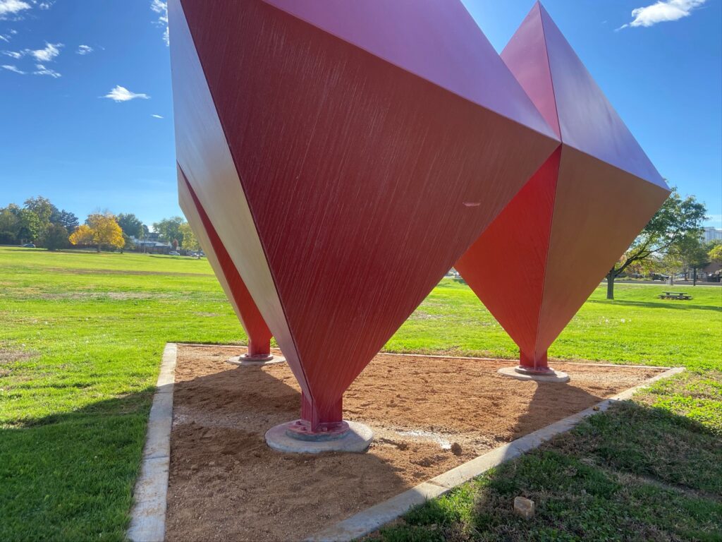 A close-up view looking up at a large geometric steel sculpture at Burns Park. The piece consists of multiple large angular diamond and triangular forms in deep red and orange-red tones, meeting at pointed bases anchored to a gravel pad. Green grass, trees, and a blue sky are visible in the background.
