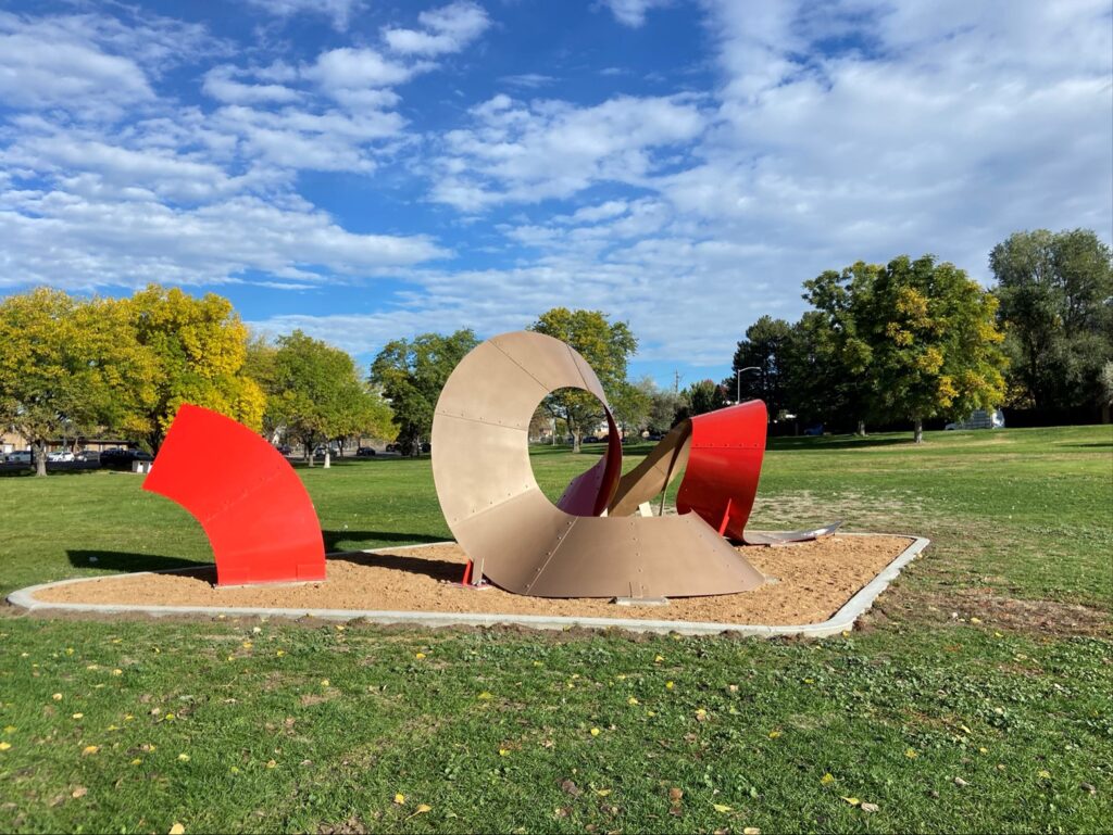 A large abstract steel sculpture in a grassy area of Burns Park on a sunny autumn day. The piece features a large tan circular loop form at the center with two bold red curved panels flanking it on either side, all set on a gravel pad. Trees with fall foliage and a blue sky with clouds are visible in the background.