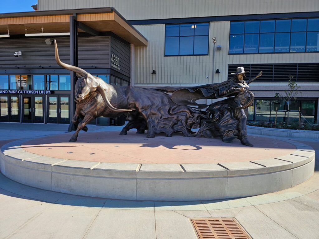 A large bronze sculpture on a circular raised platform outside the National Western Complex. The dynamic composition depicts a cowgirl in a wide-brimmed hat and flowing cape holding a lasso, facing a charging longhorn steer with dramatically sweeping horns. The figures are connected by fluid, wave-like forms suggesting movement and energy. The building entrance is visible in the background.