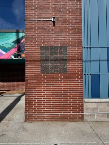 A 3x3 grid of dark bronze relief tiles mounted on a red brick exterior wall of the Ross-Phyllis Bigpond Branch Library. The tiles feature abstract and figurative imagery with textured, embossed surfaces. A colorful mural is partially visible on the left edge of the frame, and a security camera is mounted above the artwork.
