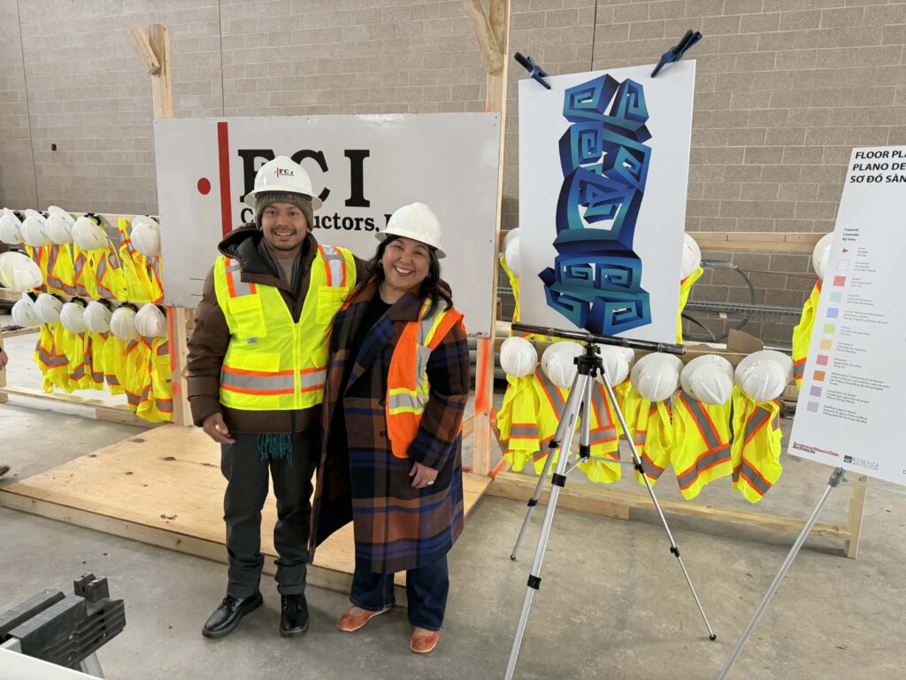 Two people wearing hard hats and safety vests smile together at an indoor construction site topping off ceremony. To their right, a rendering of a tall, stacked sculptural artwork in shades of blue with bold geometric and spiral forms is displayed on an easel. Yellow safety vests and white hard hats hang on a rack in the background alongside an FCI Constructors sign.
