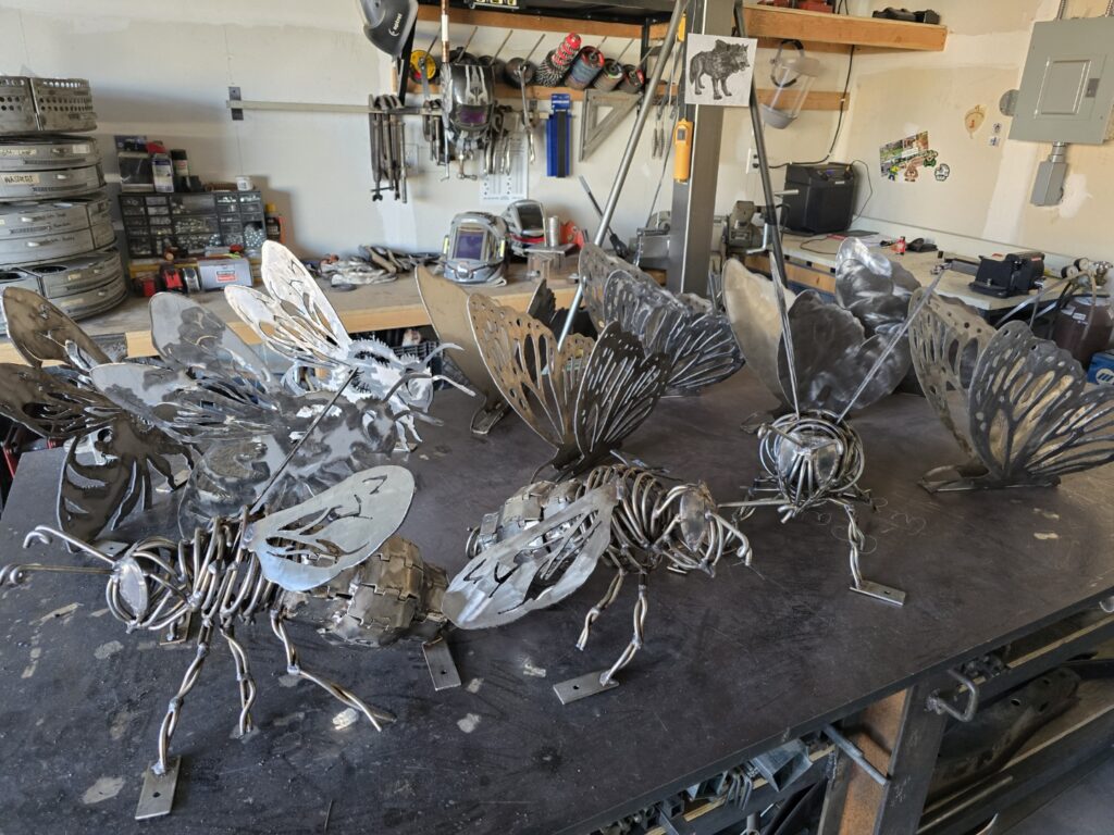 Five detailed metal sculptures of bees and butterflies in various sizes arranged on a worktable in a fabrication studio. The pieces are constructed from welded steel with intricate cutout wings and wire legs, shown in raw unfinished metal. Welding tools, helmets, storage bins, and a reference image are visible on shelves and pegboards in the background.