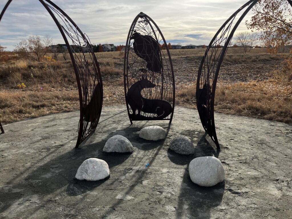 Image 1 (story circle, close-up): "A close-up view of the 'Habitats' sculpture installation at Sand Creek Regional Greenway. Three dark steel arch forms frame a circular concrete pad, with the central arch featuring a detailed silhouette of a fox and a bird amid intricate branching and botanical cutwork. Five rounded granite boulders are arranged on the pad as seating. Dry autumn grassland and residential rooftops are visible in the background under a partly cloudy sky.