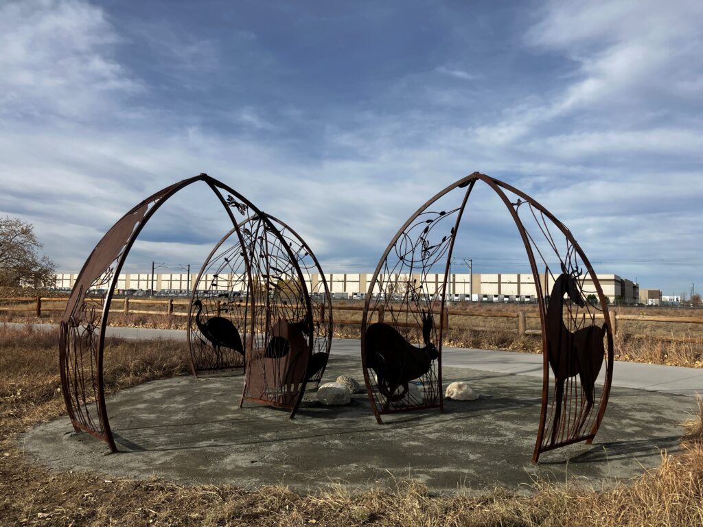 A wider view of the 'Habitats' installation showing four dark steel arch sculptures arranged in a circle on a concrete pad along the Sand Creek trail. Each arch encloses a different animal silhouette — including a heron, a bison, and a horse — set within delicate wire-like botanical framework. Small boulders sit at the center. Dry prairie grassland, a fence line, and an industrial warehouse building are visible in the background under a blue sky with clouds.