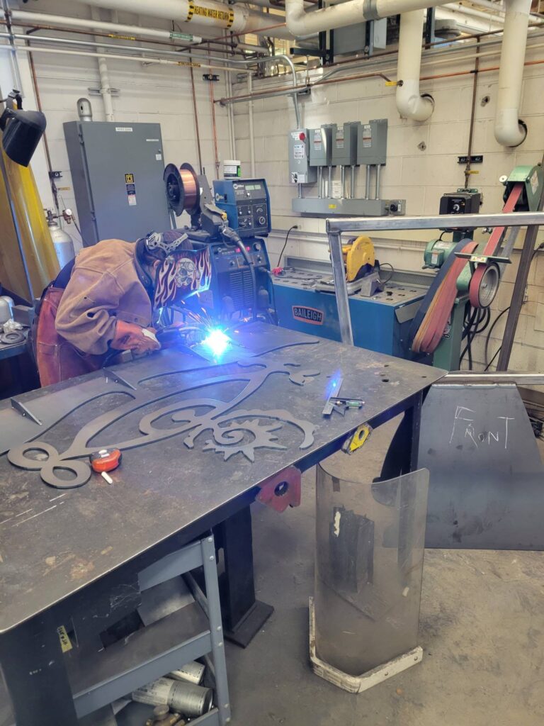 An artist in a leather welding jacket and decorative helmet welds a large flat metal cutout on a worktable in a fabrication studio. The cutout features an intricate organic design with curling and floral forms, visible on the dark steel surface. Bright blue welding sparks illuminate the work area. Industrial equipment including a welder, belt sander, and overhead pipes are visible in the background.