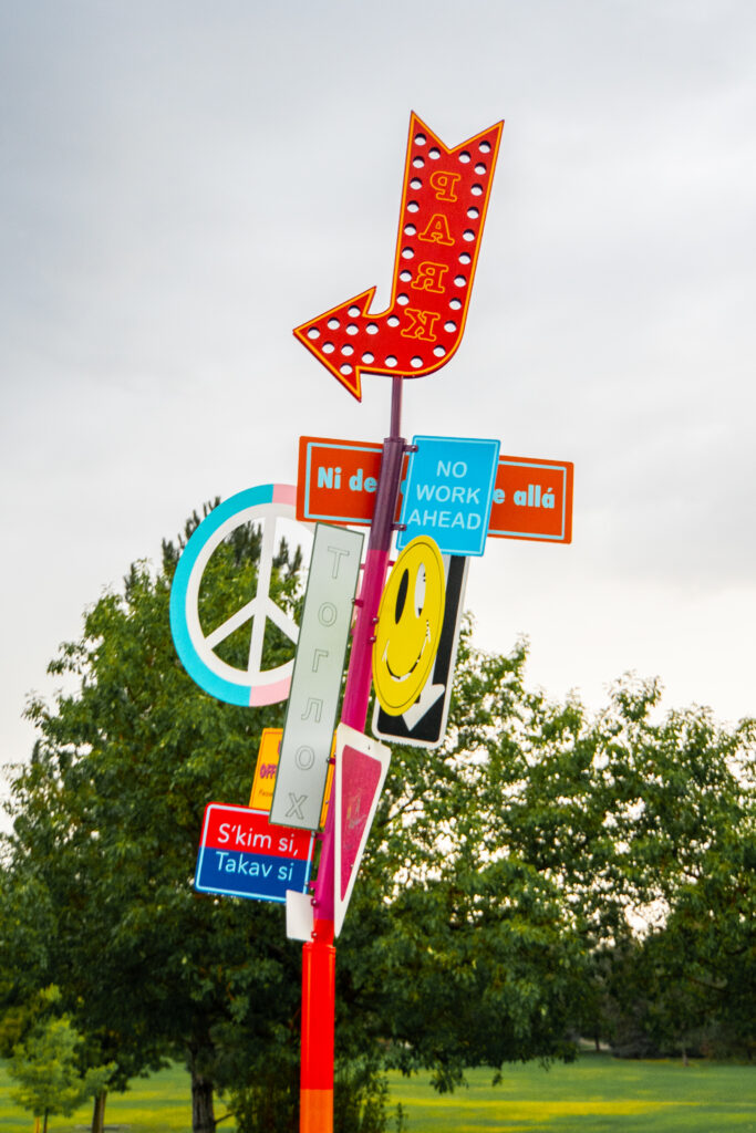A close-up of the 'Sign of the Times' sculpture showing the layered signs clustered on the pole. Visible text includes 'No Work Ahead,' 'S'kim si, Takav si,' and partial phrases in multiple languages. Other elements include a red marquee arrow with the word 'PARK' in reverse, a peace sign in teal and pink, a yellow smiley face on a black background, and a vertical sign with Cyrillic characters. Trees are visible behind.