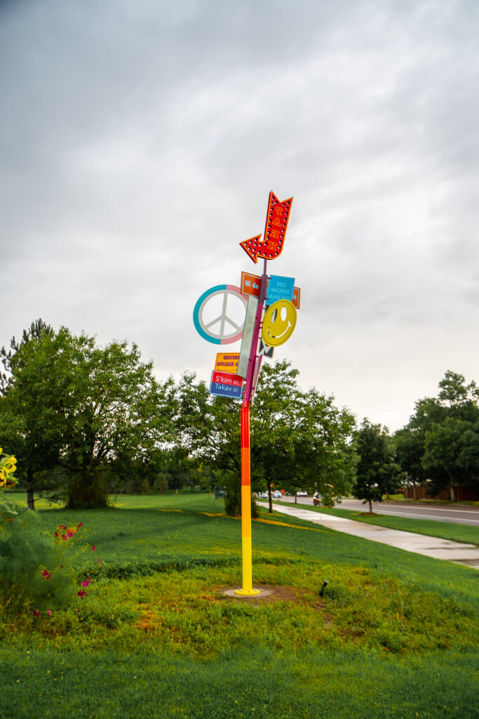 A tall, colorful sculpture pole standing in a grassy area of Bible Park. The pole is painted in stripes of yellow, orange, red, and purple, and is clustered with an eclectic mix of signs and symbols including a peace sign, a smiley face, a marquee-style arrow, and several small text signs. Trees and an overcast sky are visible in the background.