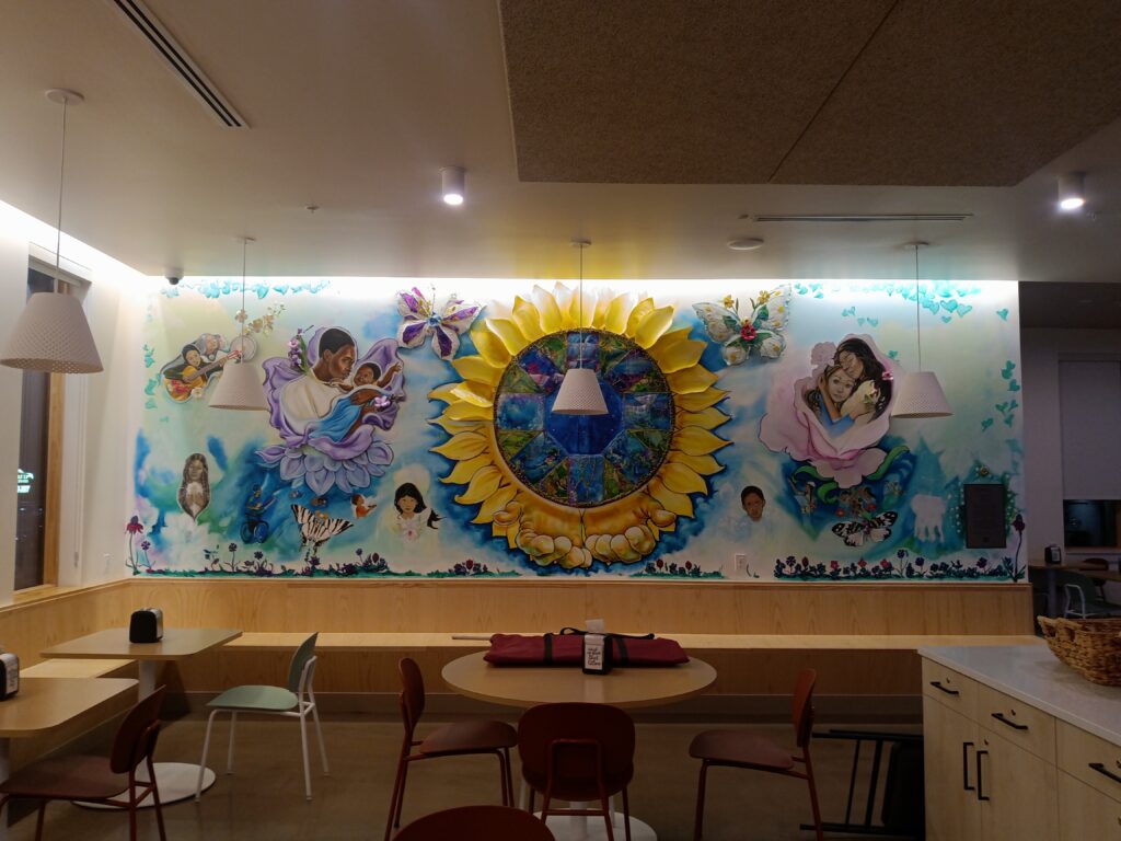 A large, colorful mural in progress covering the full length of a communal dining room wall. The composition centers on a large sunflower with a detailed circular mosaic at its center, surrounded by figures of women and children in tender embraces nestled within flowers. Butterflies, birds, and small figures are scattered throughout a soft blue sky background, with a border of wildflowers along the bottom. Three-dimensional sculptural elements including butterfly wings and flower petals extend from the wall's surface. Dining tables and chairs are visible in the foreground.