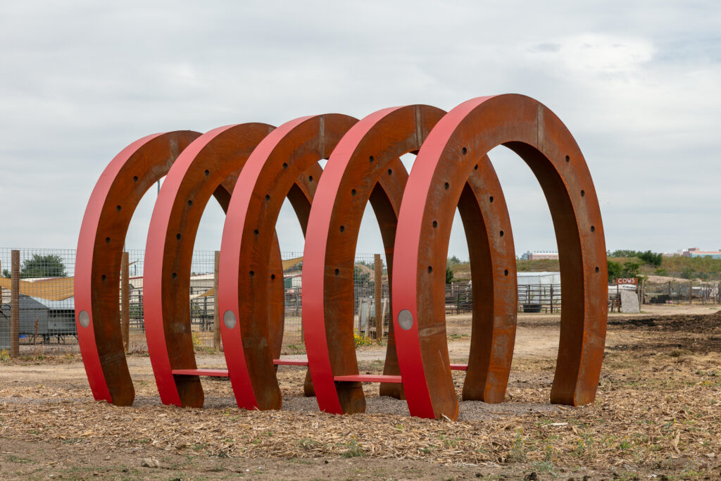 Five large horseshoe-shaped steel arches arranged in a row at The Urban Farm, forming a tunnel-like passageway. The arches have a weathered rust patina on the outer faces and are painted red on the inner edges, with circular holes punched along the curves. Red benches connect the bases. Farm structures, fencing, and an overcast sky are visible in the background.