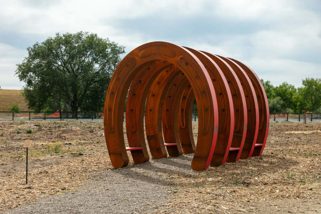 A front-facing view of the 'Talking Horseshoes' sculpture at The Urban Farm, showing the nested arches creating a tunnel effect. The innermost arch features engraved decorative detailing along its curve. A small artwork plaque on a stake is visible in the foreground, with trees and a fence line in the background under a cloudy sky.
