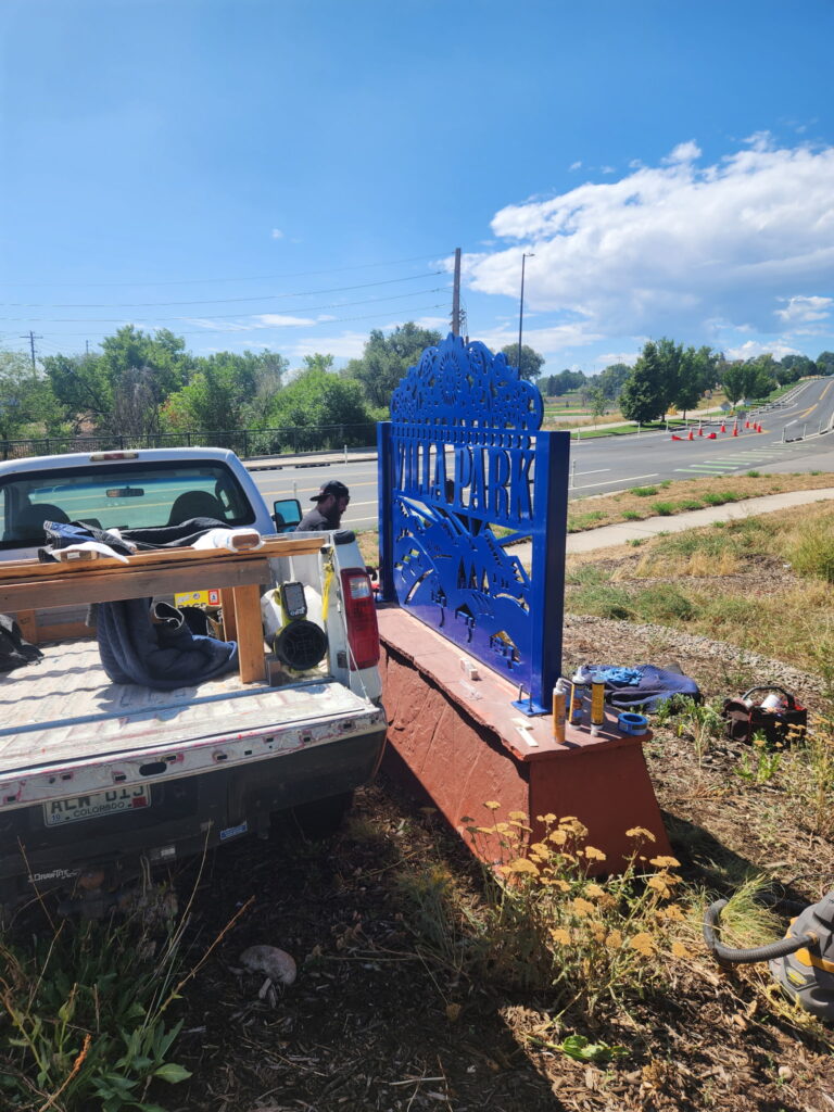 A bright cobalt blue steel gateway sign reading 'Villa Park' being reinstalled along a roadside on a sunny day. The sign features decorative cutout patterns along the top edge and landscape imagery including mountains and trees. It is mounted on a red painted concrete base. A flatbed truck and installation tools are visible in the foreground, with a worker nearby.