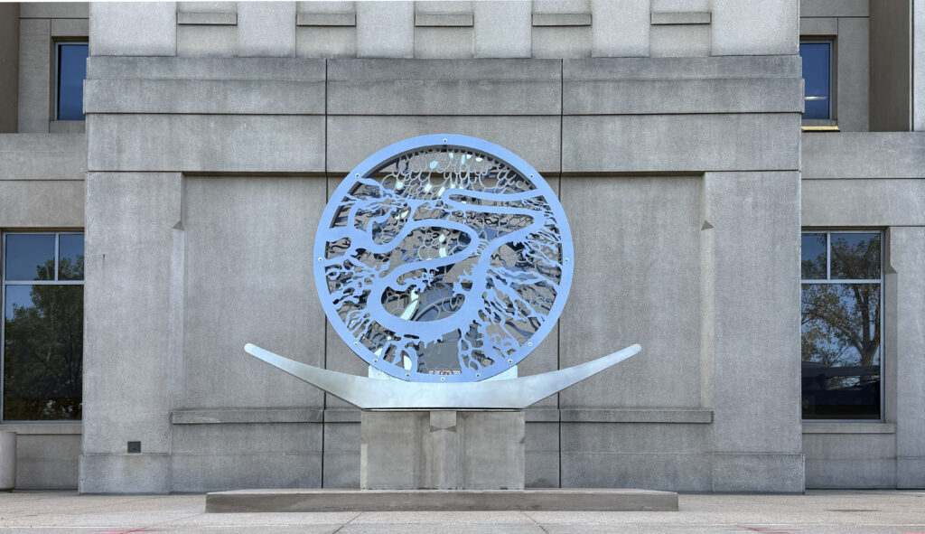 A close-up, straight-on view of the same circular steel sculpture mounted against the Wastewater Management building's stone facade. The cutout pattern of layered, fluid shapes is more visible at this angle, with the building's windows and surrounding trees reflected in the polished metal surface. The curved arms of the base extend symmetrically to either side.