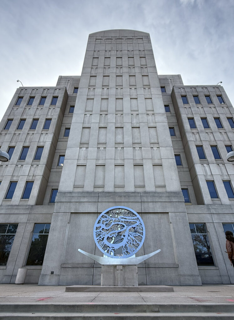 A large circular polished steel sculpture installed on a pedestal in front of a multi-story Art Deco building. The disc features an intricate cutout pattern of flowing, organic forms suggesting water or river systems. Two curved horizontal arms extend from either side of the pedestal base. The photo is taken from street level looking up, emphasizing the building's vertical limestone facade and the sculpture's scale against it.