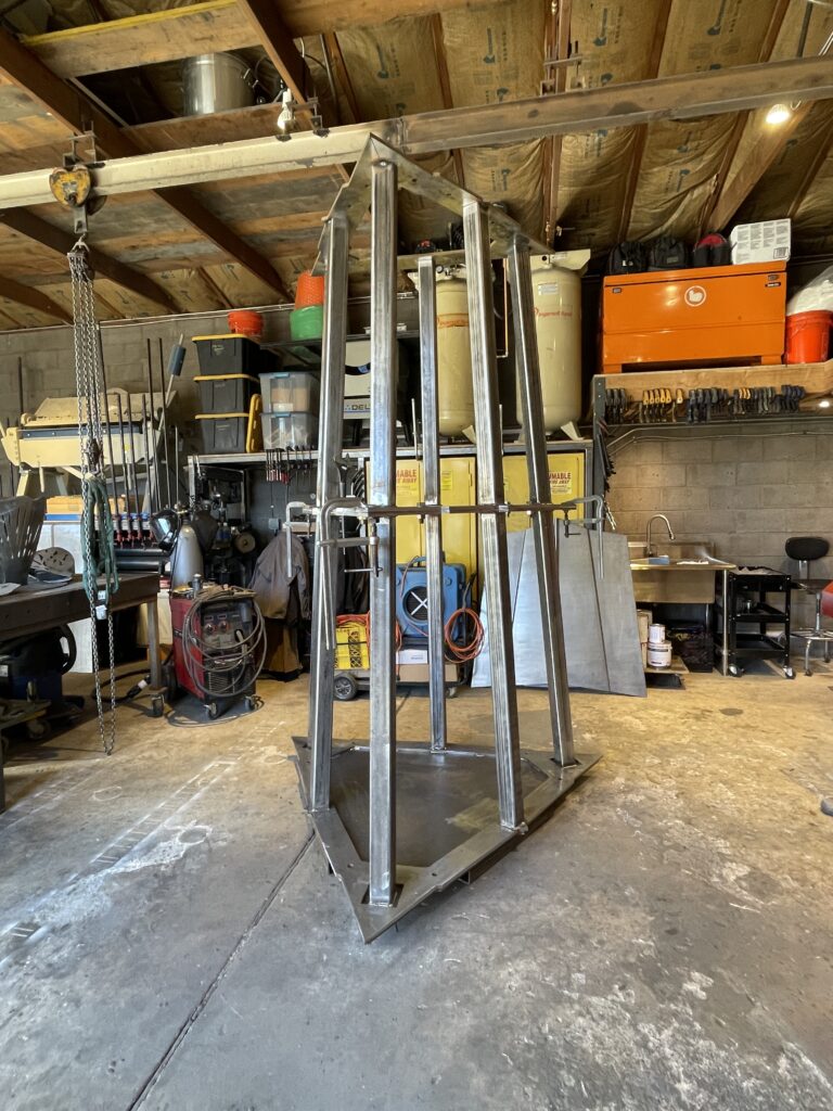 Steel sculpture components in fabrication in an artist's workshop, showing angular metal frames suspended by a chain hoist.