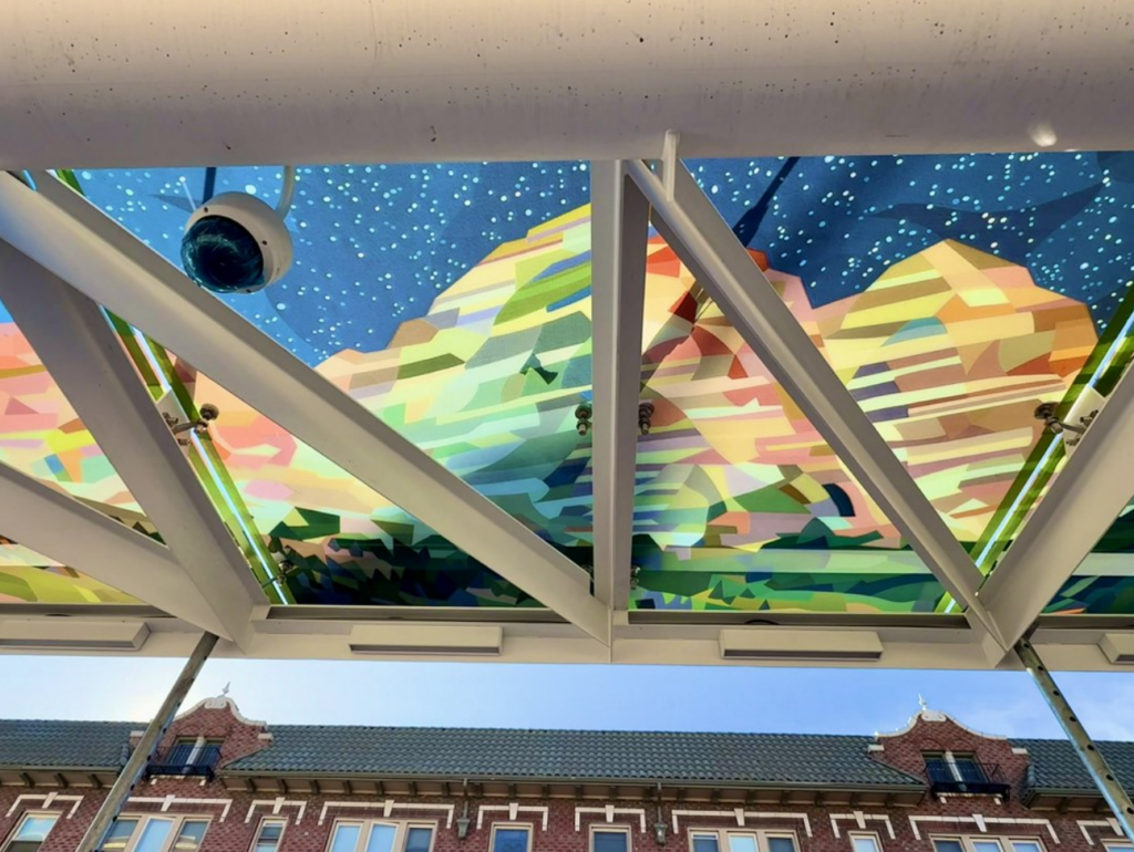 Detail of a colorful art glass panel installed on the ceiling of the westbound Pennsylvania Street Colfax BRT station, showing a vivid landscape scene viewed through the station's structural beams.