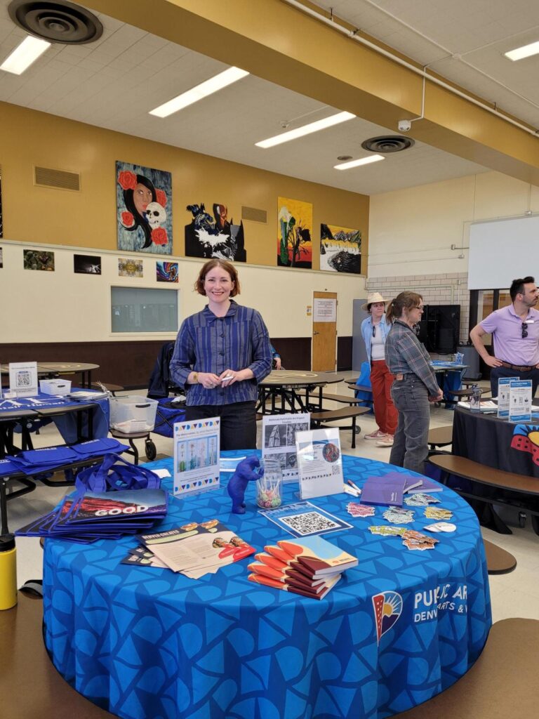 A woman with short red hair standing at a Denver Public Art information table inside a high school gymnasium during a community outreach event.