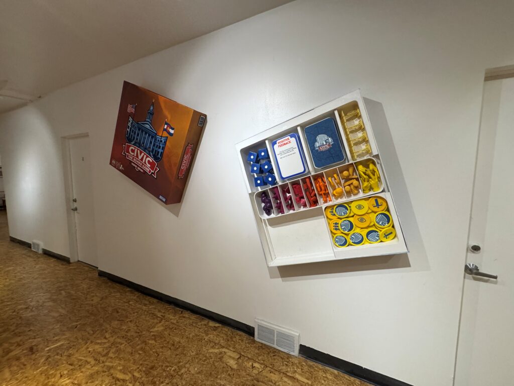 A board game-style artwork installation on a white wall, featuring a custom game box and a grid of colorful game pieces and tokens in a display case.