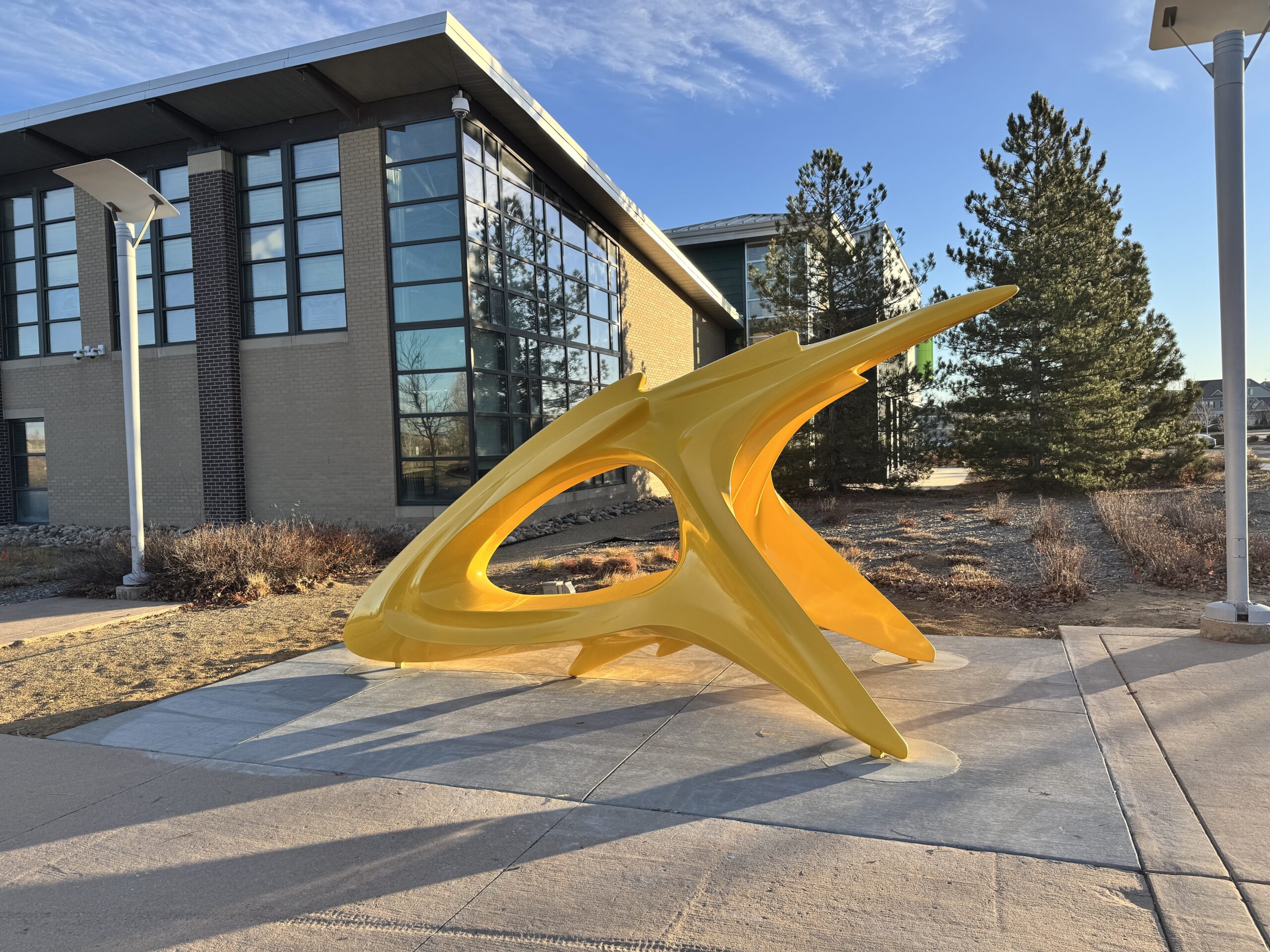 A large, glossy yellow sculpture with sweeping organic forms sits on a concrete pad outside the Green Valley Ranch Recreation Center. The piece has curved, wing-like extensions and an open oval void at its center, giving it a dynamic, almost aerodynamic quality. A brick and glass recreation center building, pine trees, and a blue sky are visible in the background.