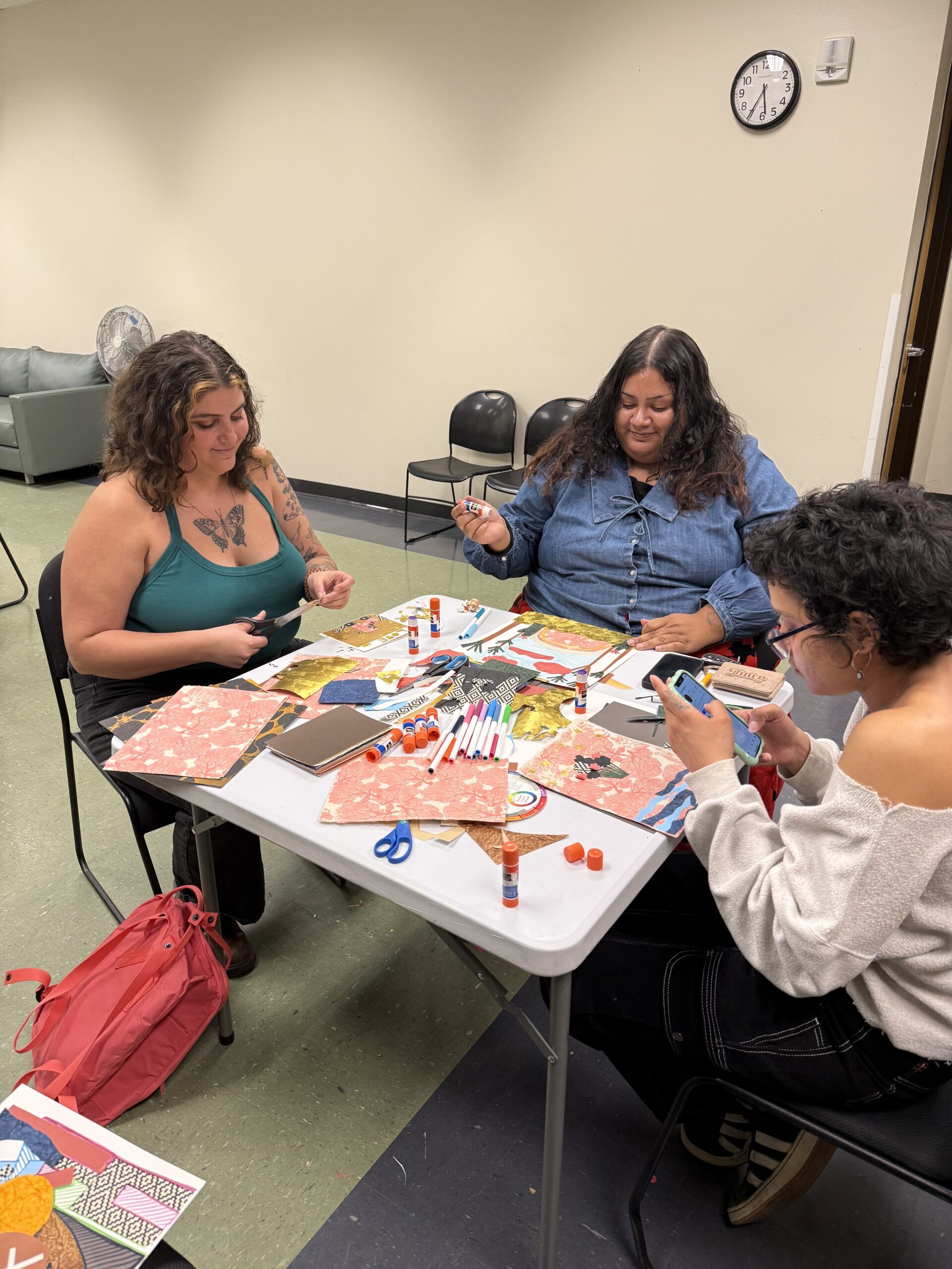 Three people sit around a folding table covered in colorful patterned paper, scissors, markers, and glue sticks, engaged in a collage-making workshop. The setting is a community room with a wall clock visible in the background.