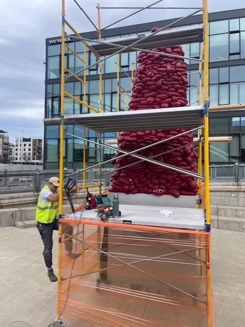 A 20-foot red fiberglass obelisk sculpture surrounded by scaffolding during maintenance work, with a pedestrian bridge visible in the background.