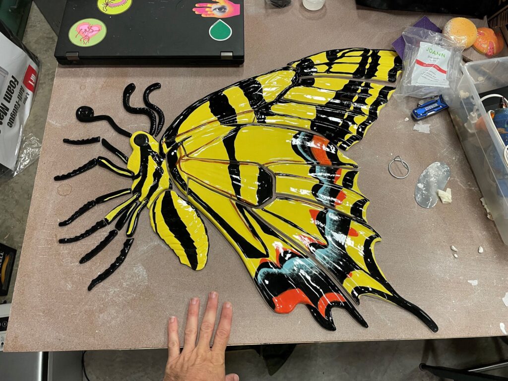 A large yellow and black swallowtail butterfly mosaic piece in progress on a workshop table, with a hand shown for scale.