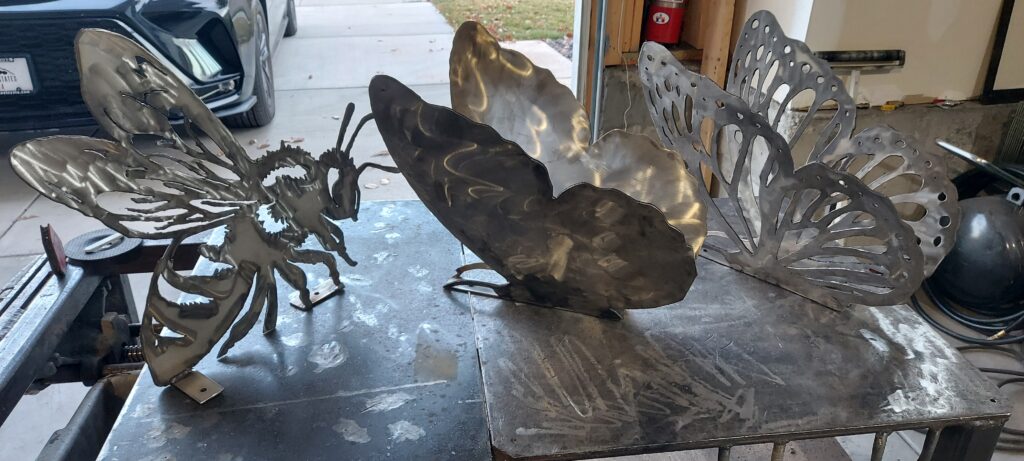 Metal butterfly and bee sculptures in various stages of fabrication laid out on a workshop table.