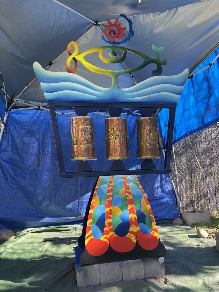 A colorful sculptural work in progress, featuring spinning copper prayer wheel cylinders beneath a painted metal totem with wave and eye motifs.