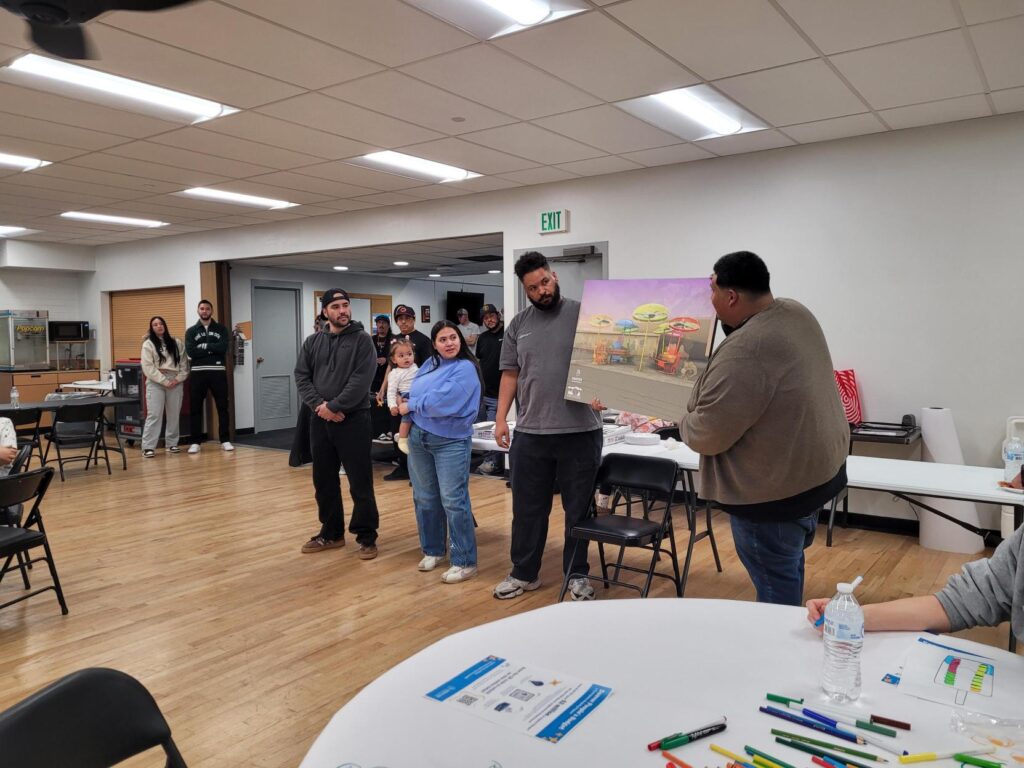 Community members gathered around tables at the Swansea Recreation Center during a public art engagement event, with a rendering of the planned artwork visible in the background.