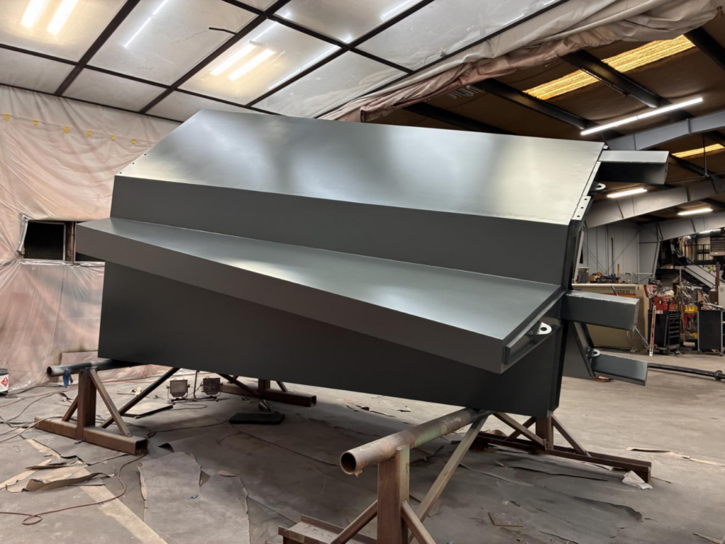 A large metal sculptural form composed of angular shapes, secured on a metal base on a fabrication floor